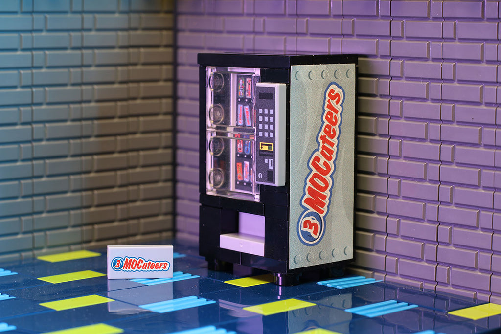 3 MOCateers - B3 Customs® Candy Bar Vending Machine made using LEGO parts displayed against a tiled wall with a custom printed 3 MOCateer card on a colorful patterned floor.