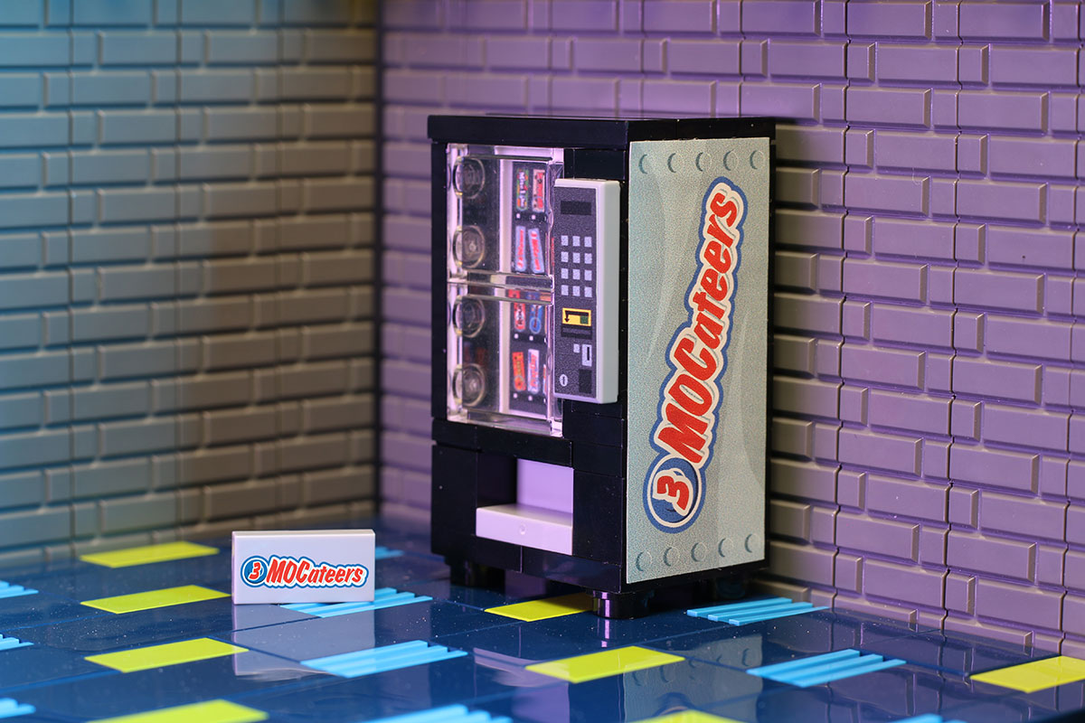 3 MOCateers - B3 Customs® Candy Bar Vending Machine made using LEGO parts displayed against a tiled wall with a custom printed 3 MOCateer card on a colorful patterned floor.