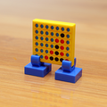 Four in a Row Printed 2x2 Tile with Stand made using LEGO parts - B3 Customs showing a miniature yellow game board with blue and red counters on blue stands on a wooden surface.