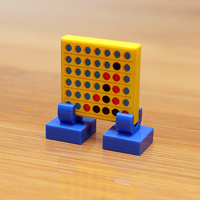 Four in a Row Printed 2x2 Tile with Stand made using LEGO parts - B3 Customs showing a miniature yellow game board with blue and red counters on blue stands on a wooden surface.