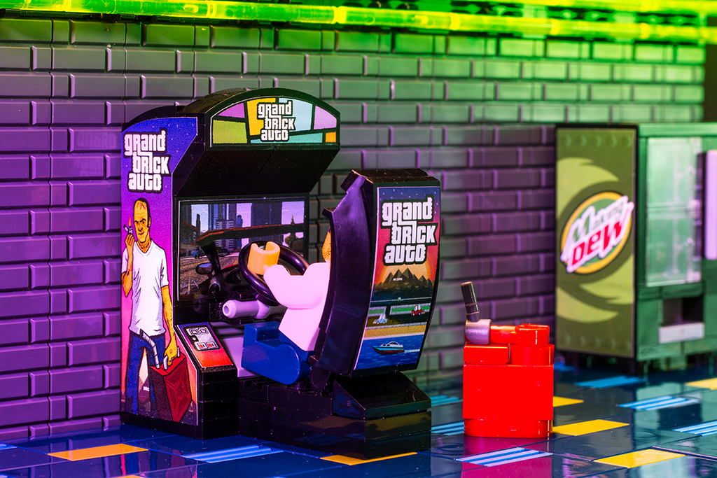 Miniature LEGO arcade racing game setup featuring a minifigure playing the Grand Brick Auto - B3 Customs Arcade Racing Game made using LEGO parts, displayed alongside a Mountain Dew vending machine.