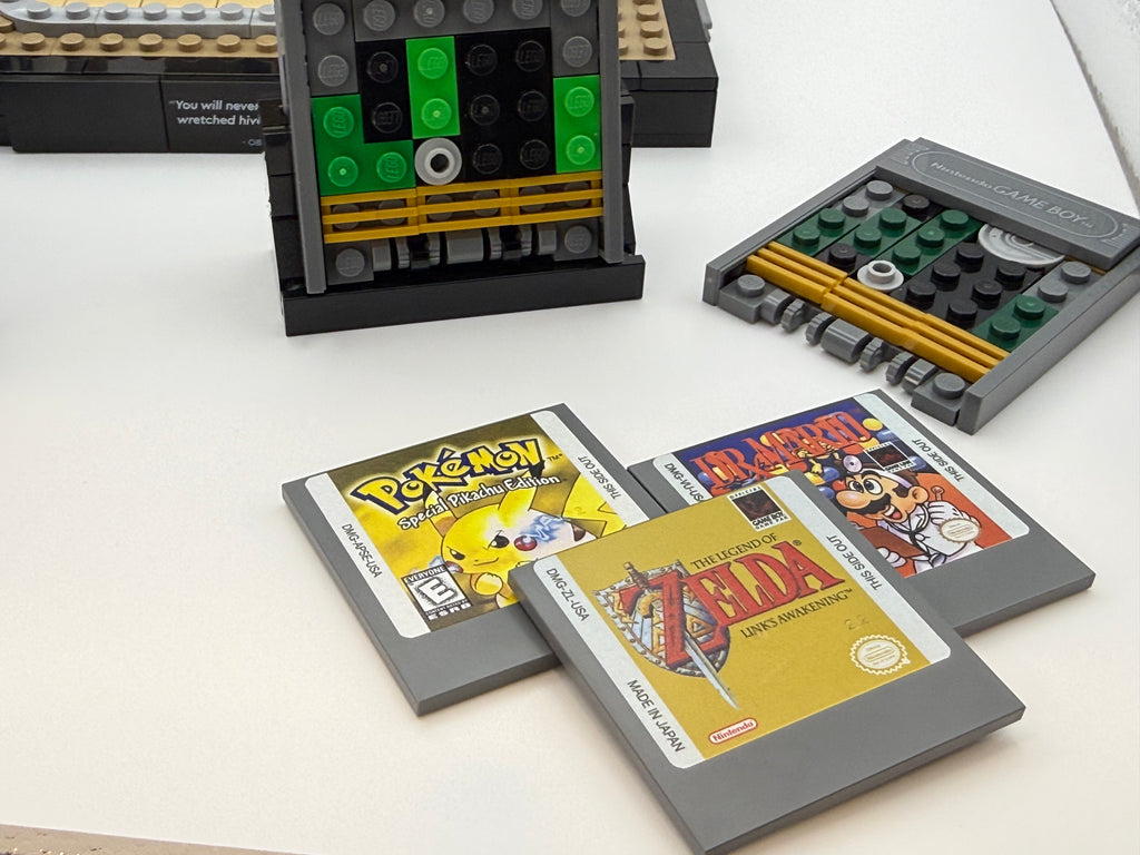 Three custom LEGO game cartridges featuring printed tiles resembling classic games like Pokémon, Mario World, and The Legend of Zelda displayed alongside brick-built game consoles using the Printed Tile Upgrade Kit for GB Set Game Cartridge.
