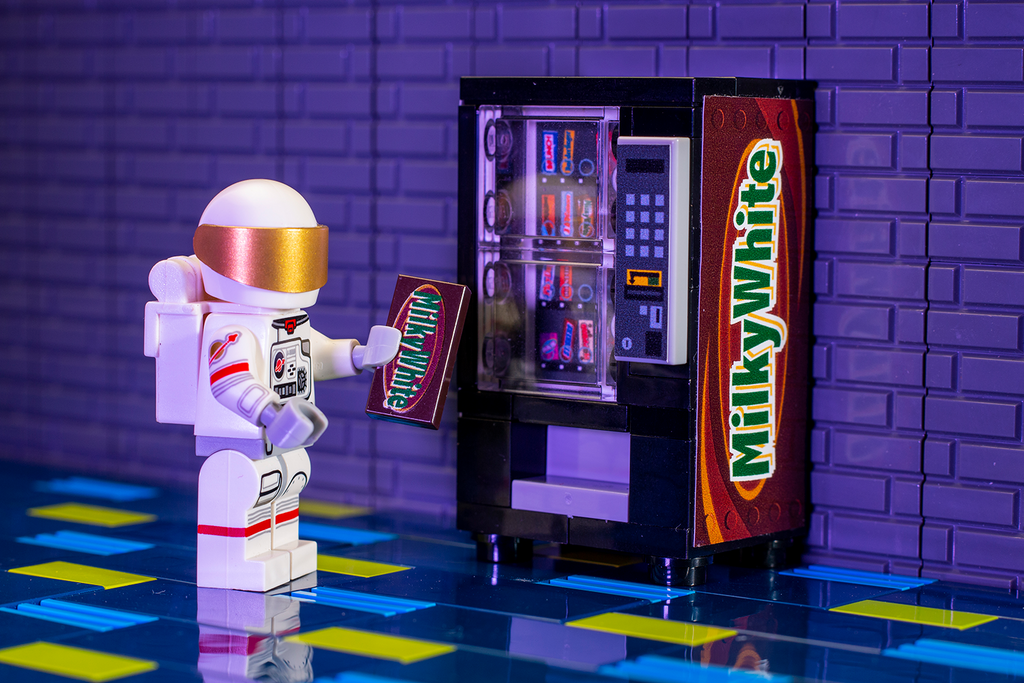 A LEGO minifigure astronaut interacts with a custom printed candy bar in front of the Milky White - B3 Customs® Candy Vending Machine, a detailed brick MOC by B3 Customs.