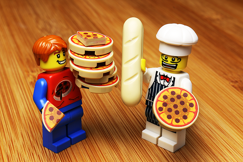 Two LEGO minifigures featuring the Pizza Pack made using LEGO parts - B3 Customs, with one holding stacked pizza slices and the other dressed as a chef holding a baguette and a whole pizza.