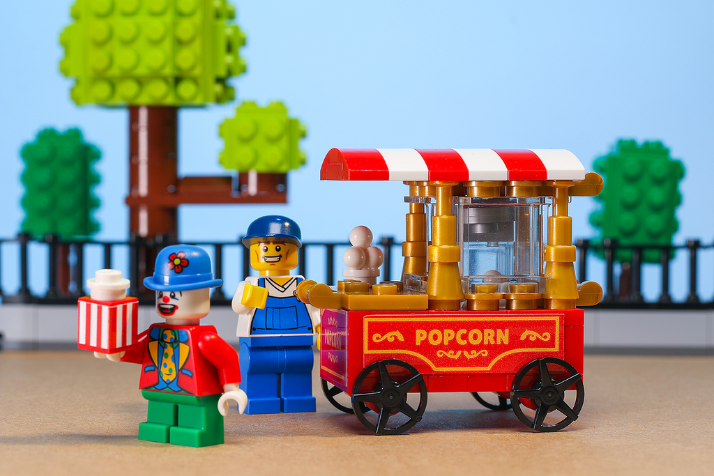 A vibrant Popcorn Vending Cart Building Set made using LEGO parts by B3 Customs, featuring a red and white striped cart with popcorn details alongside two LEGO minifigures in a playful outdoor setting.