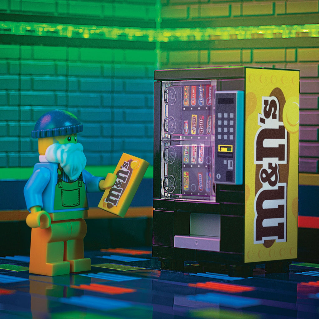 LEGO minifigure holding a peanut M&M's candy box next to a detailed M&Ns (Peanut) Candy Vending Machine made using LEGO parts - B3 Customs in a colorful, tiled setting.