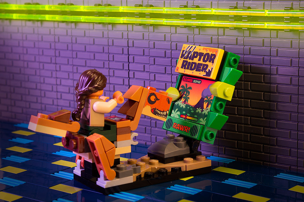 Raptor Rider - B3 Customs Arcade Game made using LEGO parts featuring a LEGO minifigure riding a dinosaur in front of the colorful arcade screen in a detailed brick-built setting.